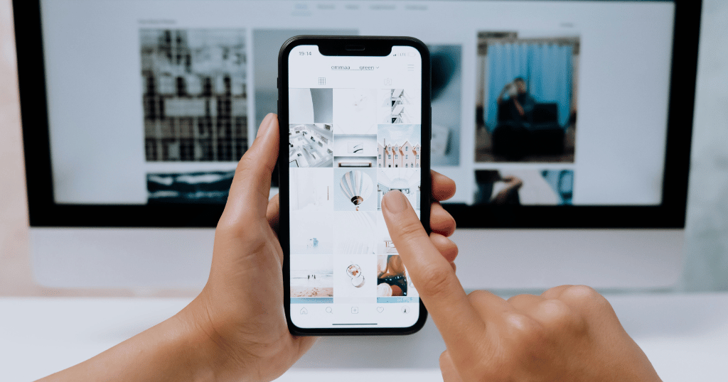 Duas mãos segurando um celular com o Instagram aberto, com uma tela de um computador no fundo