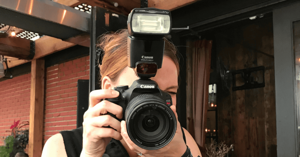 Pessoa fotografando com uma câmera DSLR Canon equipada com flash externo, destacando elementos como foco, enquadramento e composição — termos essenciais da fotografia — enquanto aponta o equipamento diretamente para a câmera.