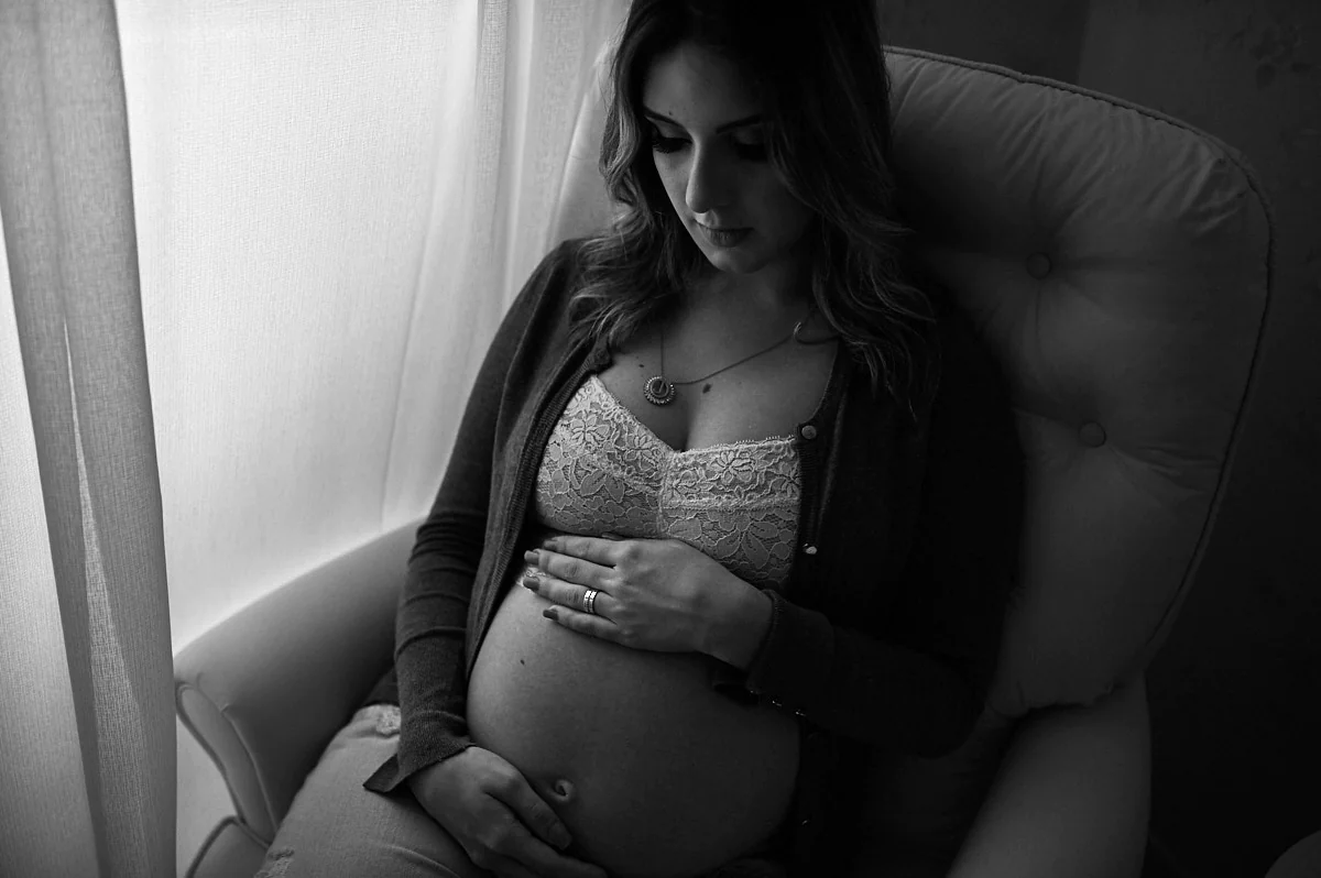 foto-de-gestante-estilo-atemporal Mulher grávida sentada, com mãos na barriga. É uma foto preto e branco