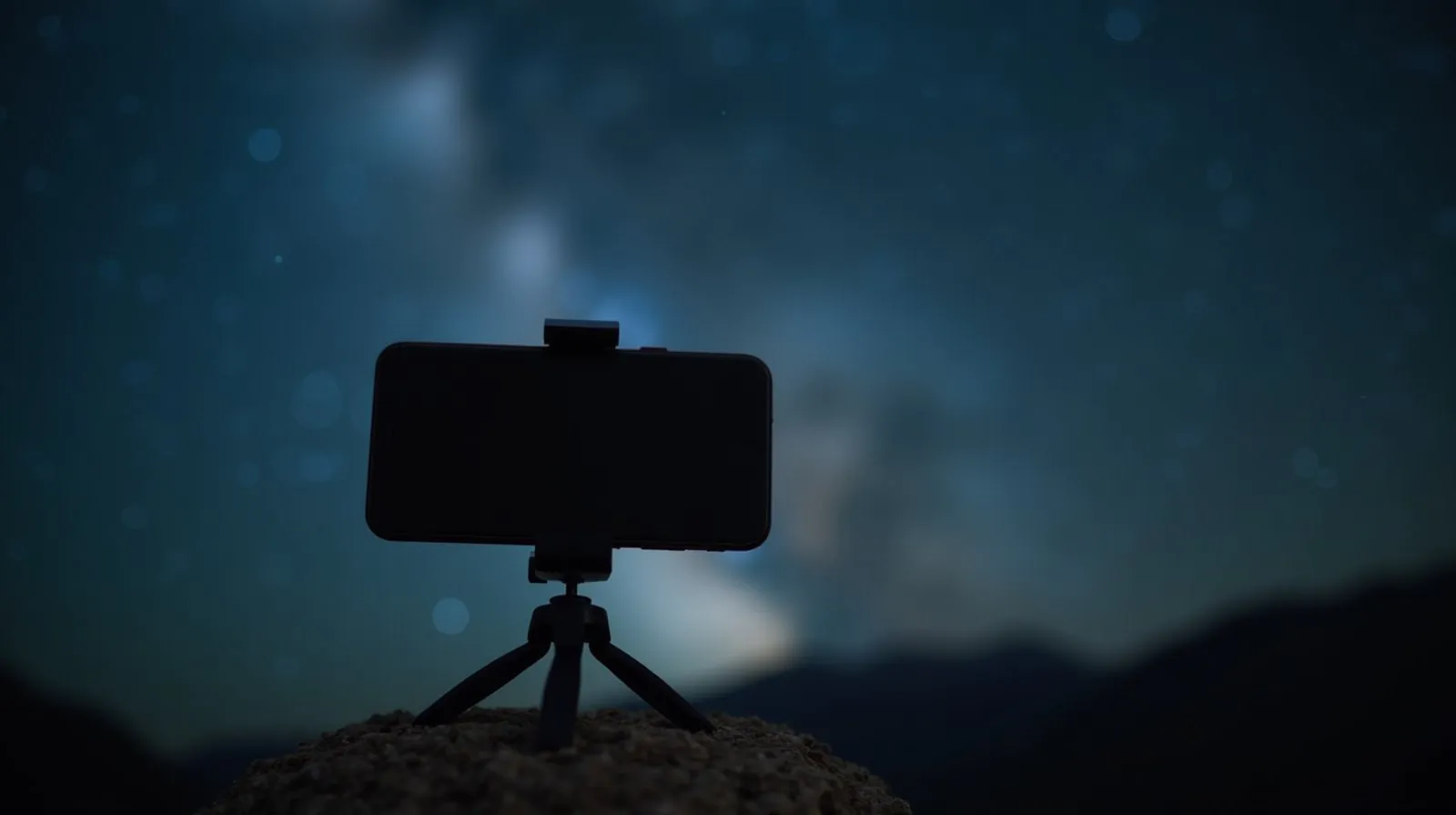 Celular preso a um mini tripé sobre uma pedra, silhueta escura contra o céu estrelado à noite